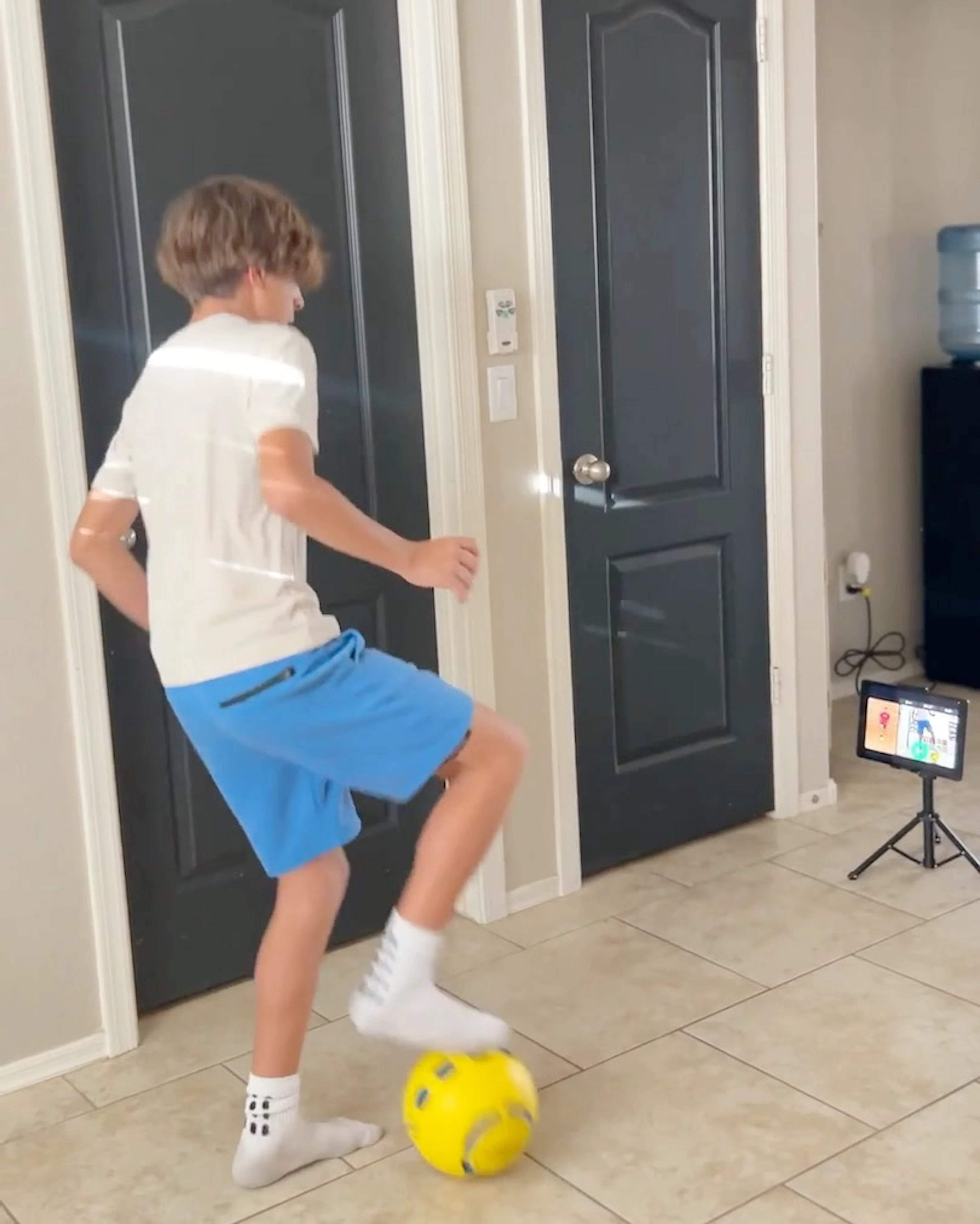 Young boy kicking a yellow Dribbleup Smart Soccer Ball indoors while following along with a tablet on a tripod