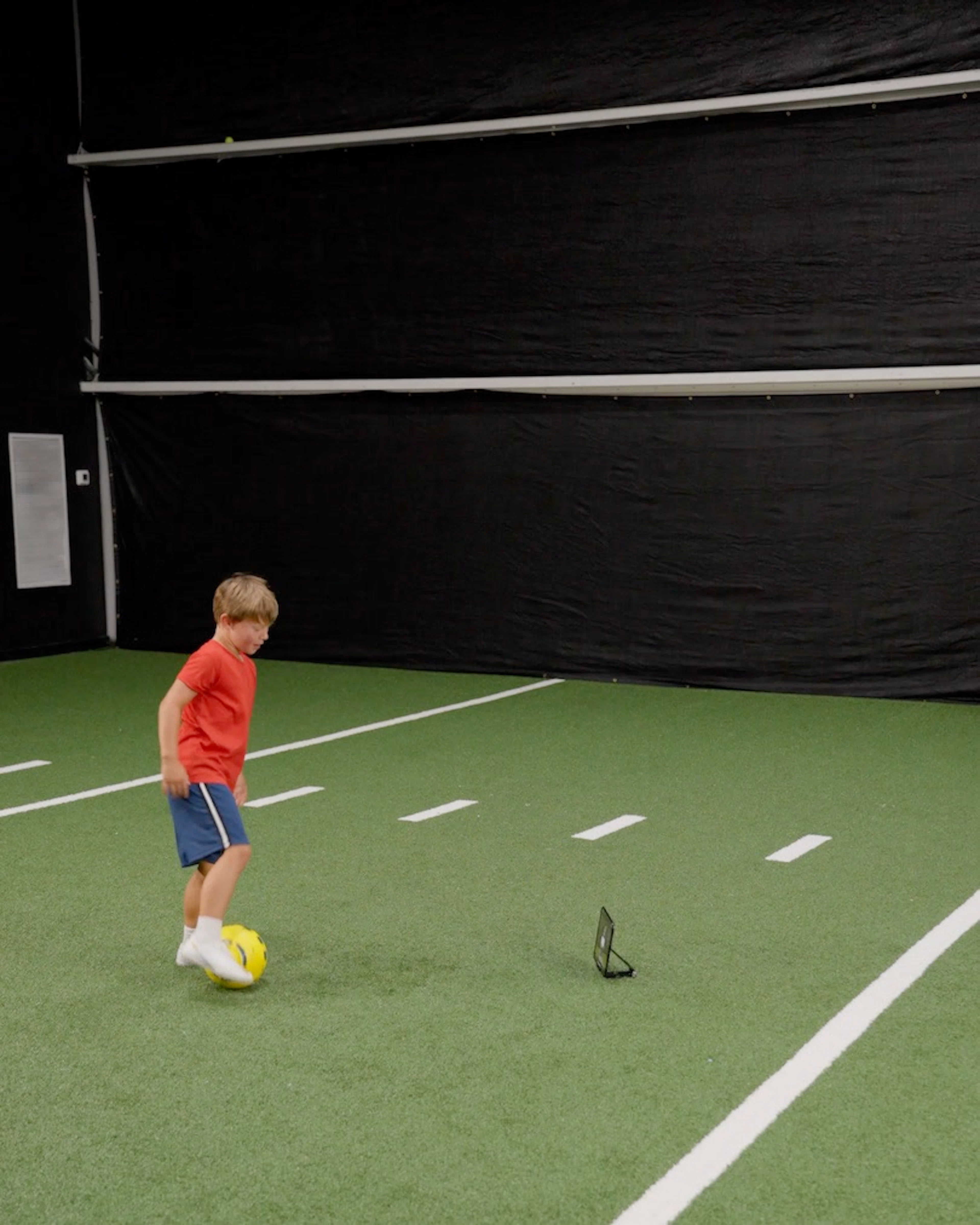 A young boy in a red shirt dribbles a yellow Dribbleup Smart Soccer Ball on indoor turf with a tablet tripod nearby