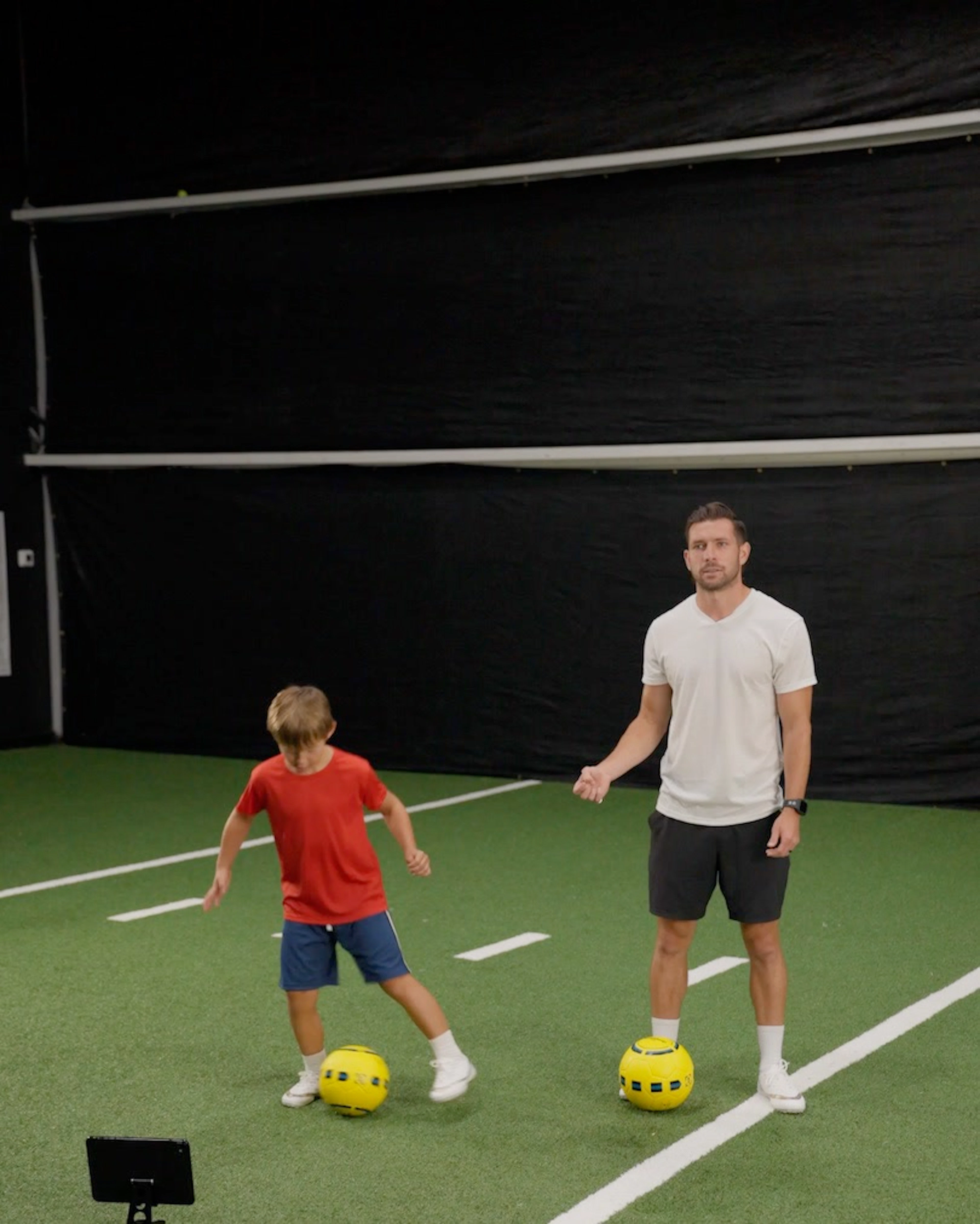 A young boy in a red shirt and an adult male practice with yellow Dribbleup Smart Soccer Balls on indoor turf with a tablet on the ground