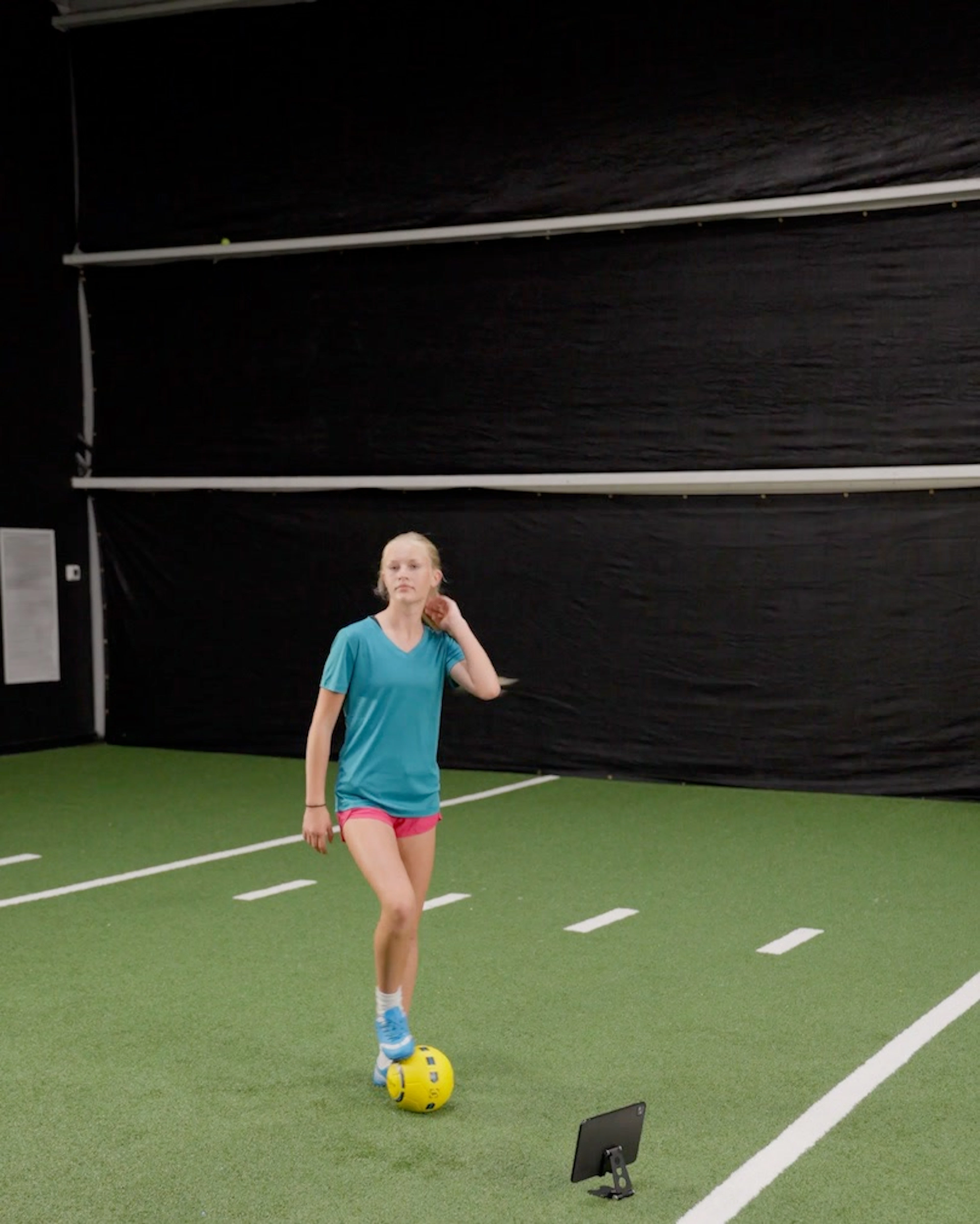 A blonde girl in a teal shirt stands with a yellow Dribbleup Smart Soccer Ball at her feet on indoor turf with a tablet nearby