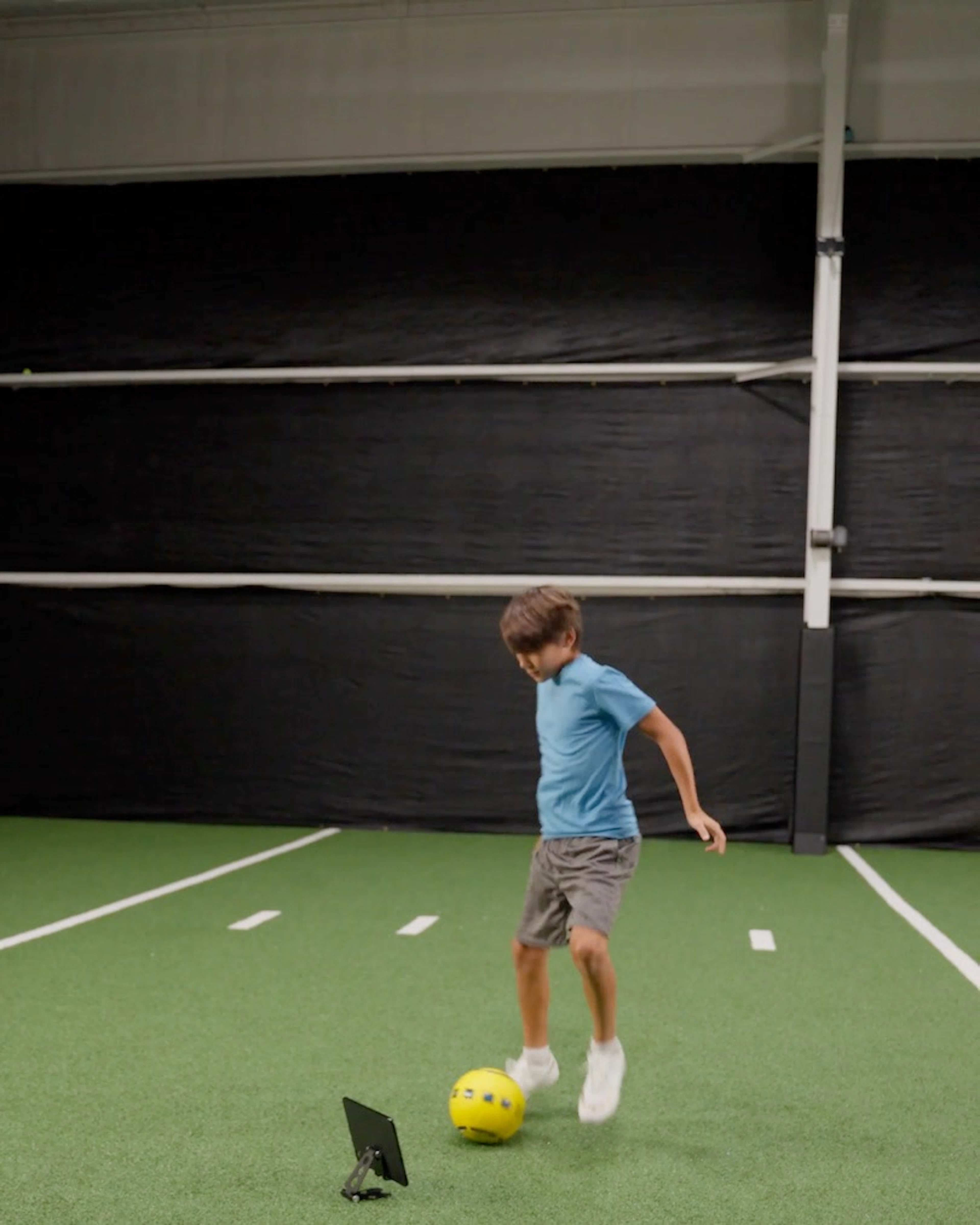 A boy in a blue shirt dribbles a yellow Dribbleup Smart Soccer Ball on indoor turf with a tablet on the ground nearby