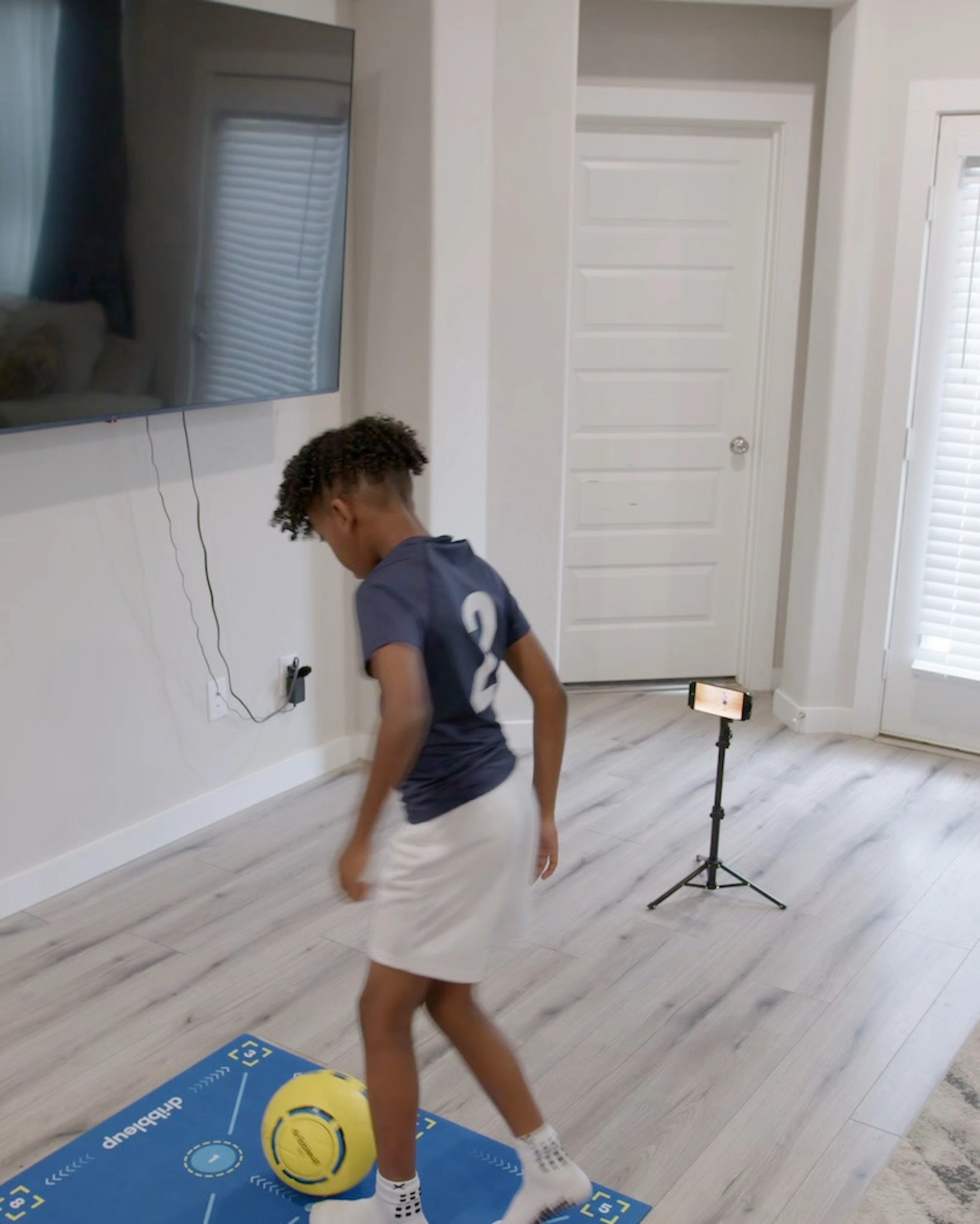 Boy dribbling a yellow Dribbleup Smart Soccer Ball on a Dribbleup mat indoors facing a phone on a tripod