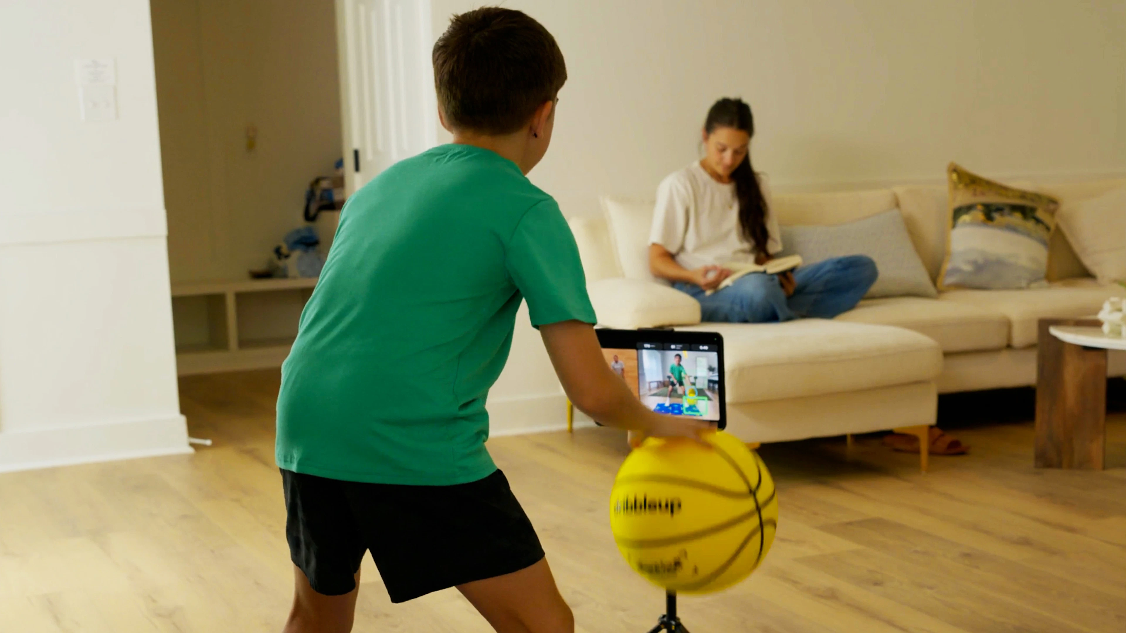 Boy dribbling the yellow Dribbleup Silent Smart Basketball in a living room while a parent reads on the couch nearby