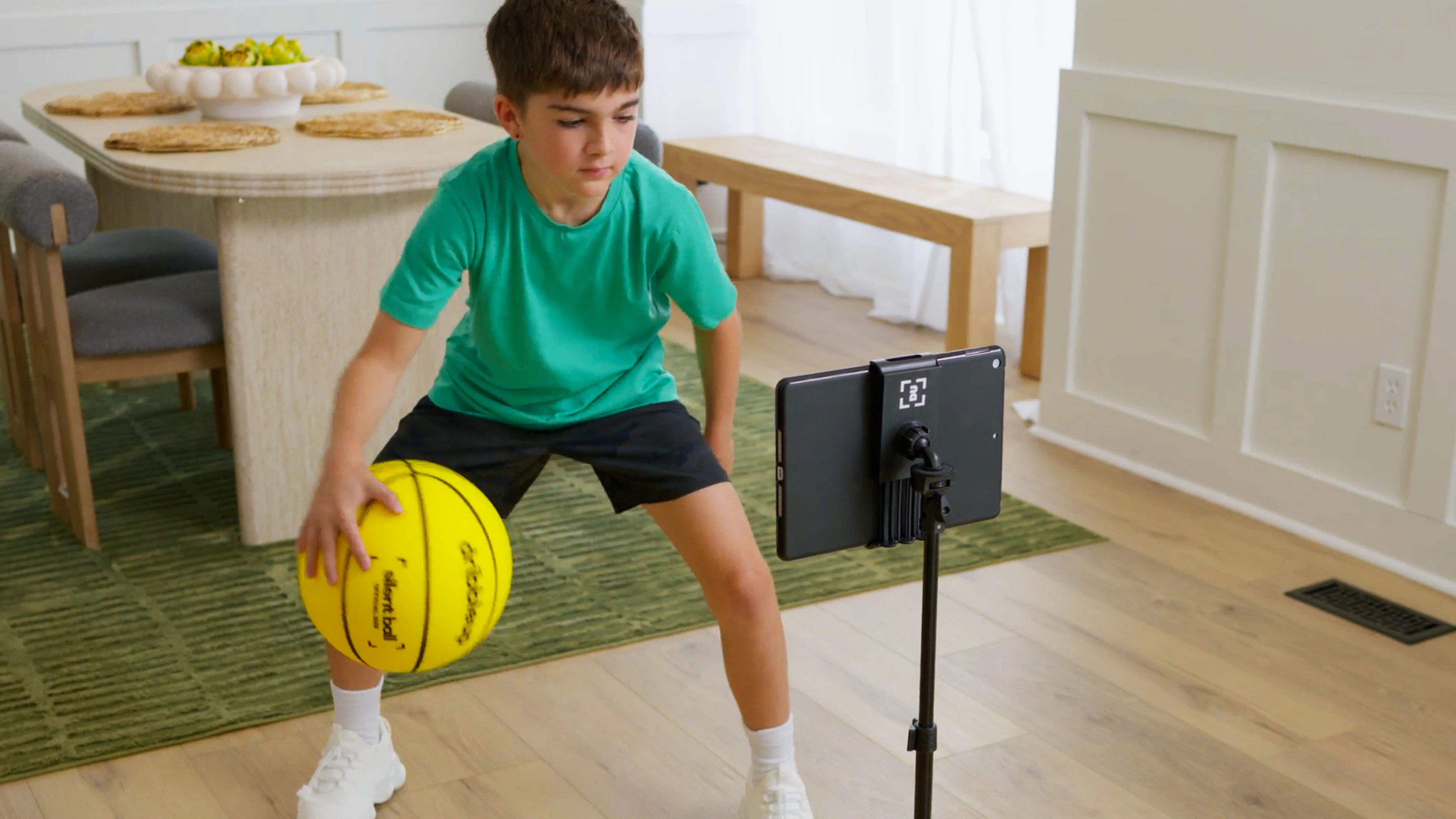 Boy running a ball-handling drill with the Silent Smart Basketball on a rug beside a dining table, following a tablet